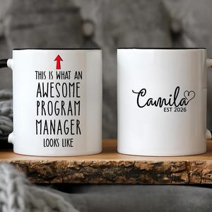 Puede incluir: Dos tazas de cerámica blancas con asas y bordes negros. Una taza dice "THIS IS WHAT AN AWESOME PROGRAM MANAGER LOOKS LIKE" con una flecha roja. La otra taza dice "Camila EST 2026" con un corazón.
