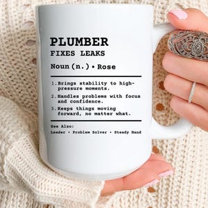 Puede incluir: Taza de cerámica blanca con el texto "PLUMBER FIXES LEAKS" en negro. La taza también incluye una definición de fontanero, enumerando sus cualidades. La taza está sostenida por una persona.