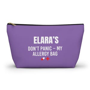 Könnte beinhalten: Ein lila Reißverschlusstasche mit dem Text "ELARA'S DON'T PANIC - MY ALLERGY BAG" in Weiß. Ein rotes Kreuz und eine weiß-rote Pillengrafik befinden sich darunter. Die Tasche hat einen goldenen Reißverschluss.