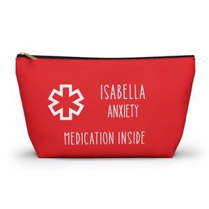 Könnte beinhalten: Rote Kosmetiktasche mit schwarzem Reißverschluss und goldenem Zipper. Der Text "ISABELLA ANXIETY MEDICATION INSIDE" ist weiß. Links befindet sich ein weißes Medizinsymbol. Für Medikamente.