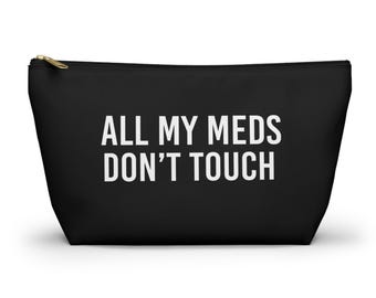 May include: A black zippered pouch featuring the white text ALL MY MEDS DON'T TOUCH centered on the front. The accessory has a gold-toned metal zipper pull at the top and a trapezoidal shape with a wide base and a narrower top edge.