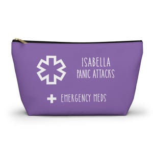 Könnte beinhalten: Ein lila Reißverschlusstasche mit weißem Text und Symbolen. Der Text lautet "ISABELLA PANIC ATTACKS" und "EMERGENCY MEDS". Auf der linken Seite der Tasche befindet sich ein weißes Lebenssternsymbol.