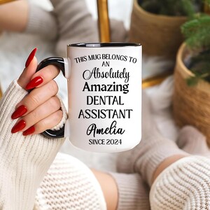 May include: White ceramic mug with black handle and rim. The mug reads "THIS MUG BELONGS TO AN Absolutely Amazing DENTAL ASSISTANT Amelia SINCE 2024" in black text. The mug is held by a person with red nail polish.