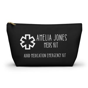 May include: Black zippered pouch with white text and a medical symbol. The text reads "AMELIA JONES MEDS KIT" and "ADHD MEDICATION EMERGENCY KIT". The pouch has a gold-colored zipper.