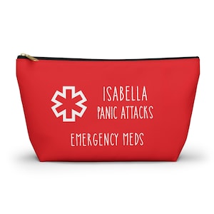 以下が含まれることがあります： 白い文字と医療シンボルが付いた赤いジッパーポーチ。「ISABELLA PANIC ATTACKS EMERGENCY MEDS」と書かれています。ポーチには黒いジッパーと金色の引き手が付いています。