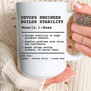May include: White ceramic mug with the text "DEVOPS ENGINEER BUILDS STABILITY" in black. The mug also includes a definition and related terms. The mug is held by a person.