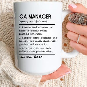 May include: White ceramic mug with the text "QA MANAGER" and a definition. The definition includes the pronunciation, responsibilities, and a humorous statement about coffee. The mug is held by a person.