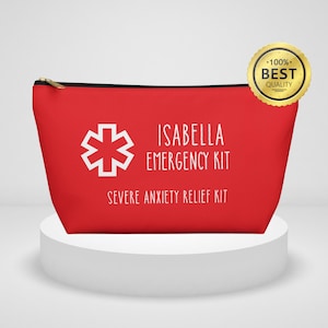 Könnte beinhalten: Rote Reißverschlusstasche mit weißem Text: "ISABELLA EMERGENCY KIT" und "SEVERE ANXIETY RELIEF KIT". Links ein weißes medizinisches Symbol. Ein goldfarbener Aufkleber mit "100% BEST QUALITY" befindet sich oben rechts.