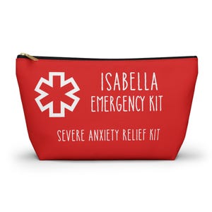 Puede incluir: Un estuche rojo con cremallera que presenta un símbolo de la Estrella de la Vida en blanco a la izquierda. El texto ISABELLA EMERGENCY KIT está impreso en blanco, seguido del texto SEVERE ANXIETY RELIEF KIT en una fuente blanca más pequeña.