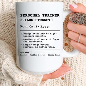 May include: White ceramic mug with the text "PERSONAL TRAINER BUILDS STRENGTH" in black. The mug also includes a definition of a personal trainer, with the word "Rose" as the noun. The mug is held by a person.