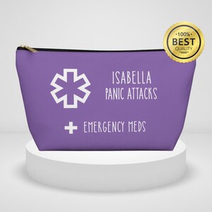 Puede incluir: Un estuche morado con cremallera con el texto "ISABELLA PANIC ATTACKS EMERGENCY MEDS" en blanco. El estuche también presenta un símbolo médico blanco y una cremallera dorada.