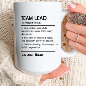 May include: White ceramic mug with the text "TEAM LEAD" in black, along with a definition. The mug also lists the responsibilities of a team lead. The text "See Also: Rose" is at the bottom of the mug.