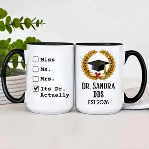 May include: White ceramic mug with a black handle and rim. One side reads "Miss, Ms., Mrs., Its Dr. Actually." The other side features a graduation cap, diploma, and the text "Dr. Sandra DDS EST 2026" within a gold laurel wreath.