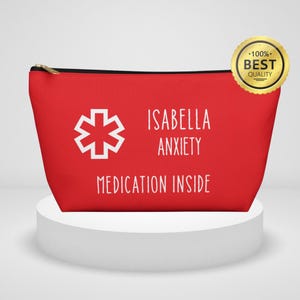 Op de afbeelding: Een rode etui met zwarte rits en witte tekst "ISABELLA ANXIETY MEDICATION INSIDE". Links staat een wit medisch symbool. Een gouden "BEST QUALITY" zegel staat rechtsboven. De etui staat op een witte sokkel.