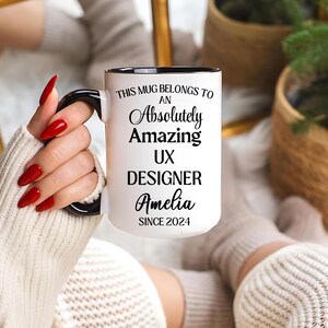 May include: White ceramic mug with a black handle and rim. The mug has the text "THIS MUG BELONGS TO AN Absolutely Amazing UX DESIGNER Amelia SINCE 2024". The mug is held by a person with red nail polish.