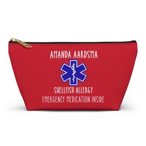 Può includere: Pochette rossa con cerniera dorata. La pochette presenta il nome "AMANDA AARDSMA" sopra un simbolo medico blu e il testo "SHELLFISH ALLERGY EMERGENCY MEDICATION INSIDE" in bianco.