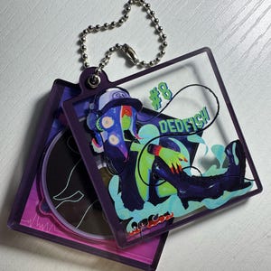 May include: A clear acrylic keychain with a purple border, featuring a colorful illustration of a character with the text "#8 DEDFISH". The keychain is attached to a silver ball chain.