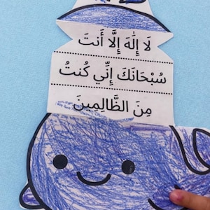 Peut inclure: Un artisanat en forme de baleine bleue avec un visage souriant et du texte en arabe. La baleine est colorée au crayon bleu et a un contour noir. Le texte est imprimé sur une section de papier blanc au-dessus de la baleine.
