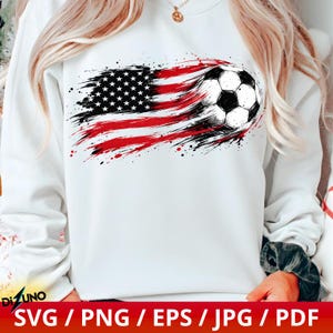May include: A white long-sleeve sweatshirt featuring a graphic print of a football integrated into a distressed American flag design. The flag pattern uses red, white, and black brushstroke effects that trail behind the football.