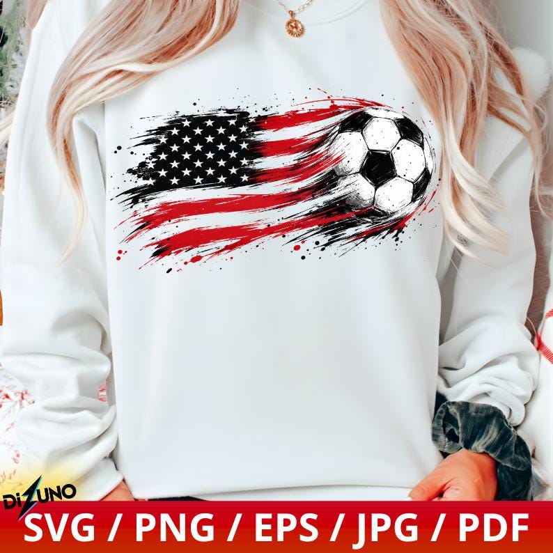 May include: A white long-sleeve sweatshirt featuring a graphic print of a football integrated into a distressed American flag design. The flag pattern uses red, white, and black brushstroke effects that trail behind the football.