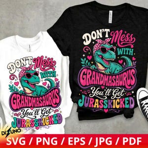 May include: A white and a black t-shirt featuring a graphic of a green dinosaur wearing pink sunglasses and a polka dot headscarf. The text reads: Don't Mess With Grandmasaurus You'll Get Jurasskicked. The design includes pink hearts and leaves.