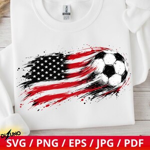 May include: A white crewneck sweatshirt featuring a graphic print of a football integrated with a stylised American flag design. The flag appears in a distressed, brushstroke style with red, white, and black ink splatters across the front.
