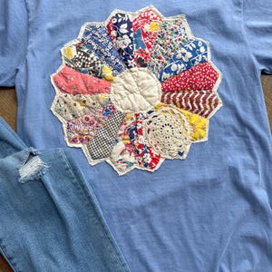 May include: A light blue t-shirt with a large, colorful, patchwork flower design on the front. The flower is made of various patterned fabrics and a crocheted center. Light blue denim jeans are visible in the lower left corner.