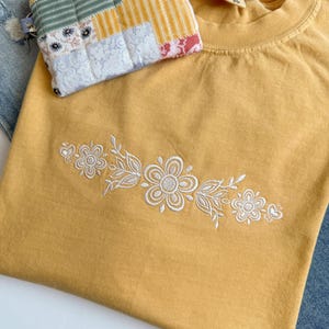 May include: Mustard yellow t-shirt with white floral embroidery. A small, patchwork fabric coin purse rests on the shirt. The purse features various fabric patterns in shades of yellow, blue, and red. The shirt is folded, and denim jeans are visible in the background.