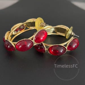 May include: A pair of gold-toned hoop earrings with oval, deep red gemstones. The earrings have a classic design with a polished finish. The gemstones are evenly spaced around the hoops, creating a bold and elegant look.