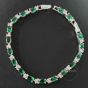 Könnte beinhalten: Ein silbernes Armband mit ovalen, smaragdgrünen Edelsteinen und kleinen, klaren Akzentsteinen. Das Armband hat eine silberne Metalleinfassung und einen Verschluss. Die Edelsteine sind gleichmäßig über die Länge des Armbands verteilt.