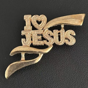 Puede incluir: Un broche de metal en tono dorado con el texto I LOVE JESUS, donde la palabra LOVE está representada por un símbolo de corazón. El diseño incluye acentos estilizados en forma de cinta en la parte superior e inferior, con acabado texturizado.