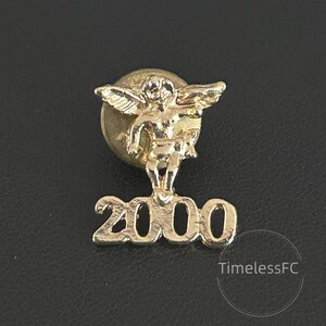 Puede incluir: Pin de metal dorado con un querubín alado sobre el número "2000". El pin tiene un diseño clásico y es probablemente un artículo conmemorativo. El pin está sobre un fondo oscuro.