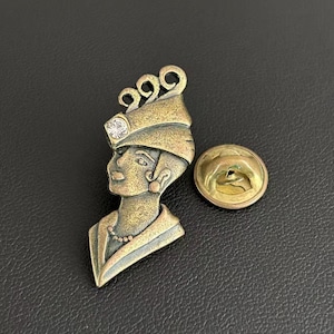 May include: A brass-toned metal brooch shaped as a profile of a person wearing a headpiece with a single clear rhinestone accent. The pin features a textured finish and includes a separate circular metal backing piece with a clutch fastener.
