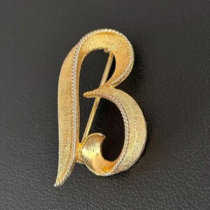 May include: A gold-tone metal brooch shaped like the letter B. The surface features a textured, cross-hatched finish with a twisted rope-style border along the edges. The design is open and fluid, with a pin mechanism visible behind the letter.
