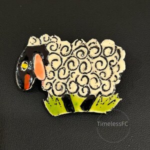 May include: A whimsical sheep brooch with a black head, a cream-colored body with black swirls, and a green base with black stripes. The sheep has a red mouth and a yellow eye. The brooch is a unique accessory.