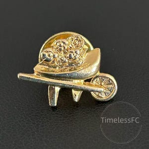 May include: A gold-toned pin featuring a wheelbarrow filled with flowers. The wheelbarrow has a detailed design, including a wheel and handles. The pin is set against a dark background.