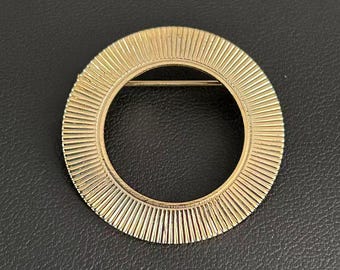 Broche circular vintage en tono dorado con diseño de rayos de sol y alfiler redondo acanalado.