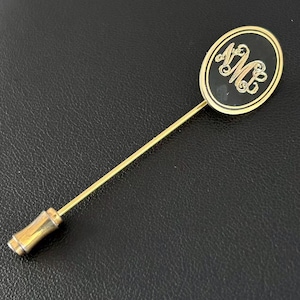 May include: Gold-toned stick pin with an oval black enamel head. The head features a gold monogram of the letters "AM" in a decorative script. The pin has a long, slender shaft and a decorative end.