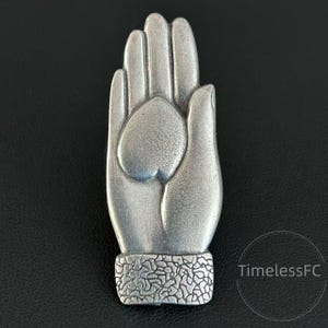 Puede incluir: Colgante plateado en forma de mano con un corazón en la palma. El colgante tiene una pulsera texturizada y se muestra sobre un fondo negro. El diseño es detallado y tiene una estética vintage.