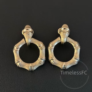 May include: A pair of gold-colored hoop earrings with a bamboo-style design. Each earring features a textured, hexagonal hoop and a decorative top piece. The earrings are displayed on a dark surface.