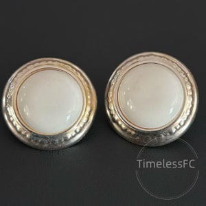 May include: A pair of circular stud earrings with a silver-coloured finish. Each earring has a large, white, circular centre stone, framed by a textured silver border. The earrings are shown against a dark background.