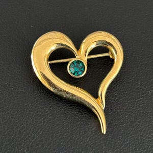 May include: A gold-toned heart-shaped brooch with a central emerald-green gemstone. The brooch has a pin closure on the back. The heart design is smooth and polished, with a slight textured effect on the surface.