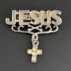 Puede incluir: Un broche de metal plateado con la palabra JESUS en una fuente de letras de bloque texturizadas. Debajo del texto hay un marco decorativo con volutas y un dije de cruz colgante. La cruz está engastada con cinco pequeños brillantes claros.