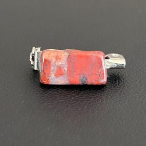 May include: A rectangular pendant with a red and tan stone, accented with black and gray inclusions. The stone is mounted with silver-colored metal findings. The pendant is displayed against a dark background.