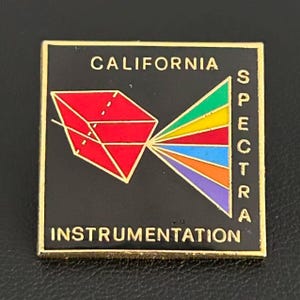 May include: A square black enamel pin with gold trim featuring the text CALIFORNIA SPECTRA INSTRUMENTATION. The design includes a red geometric prism on the left and a multi-colored light spectrum fan on the right in green, yellow, red, blue, orange, and purple.