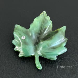 May include: Green leaf-shaped brooch with a small, sparkling crystal accent. The brooch has a glossy finish and a dark green center, with lighter green edges. The leaf design is detailed and realistic, with visible veins. A unique piece of jewelry.