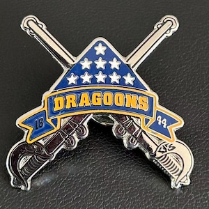 Puede incluir: Un pin plateado con dos sables cruzados. Una pancarta azul con la palabra "DRAGOONS" en amarillo cruza el centro. Encima de la pancarta hay un triángulo azul con estrellas blancas. Los números "18" y "44" están en la pancarta.