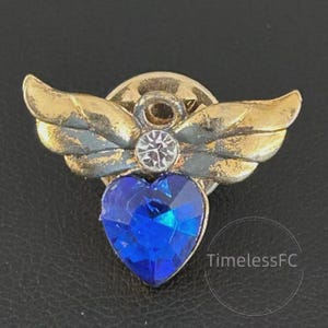 Könnte beinhalten: Eine goldfarbene Engelsnadel mit ausgebreiteten Flügeln und einem herzförmigen blauen Edelstein. Die Nadel hat einen zentralen klaren Kristall und den Text "TimelessFC" in einem kreisförmigen Design.