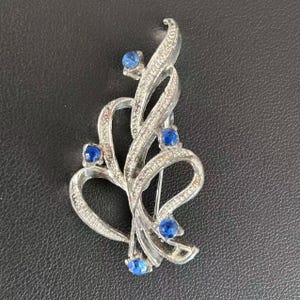 Puede incluir: Un broche de metal plateado con un diseño de cinta en espiral y superficie texturizada. La pieza está decorada con cinco pequeñas piedras redondas de color azul oscuro, colocadas a intervalos a lo largo de los bucles metálicos curvos.