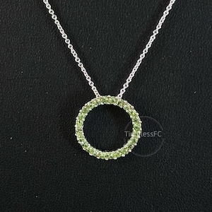 May include: A silver necklace with a circular pendant. The pendant is encrusted with small, light green gemstones. The chain is delicate and silver. The background is a solid black.